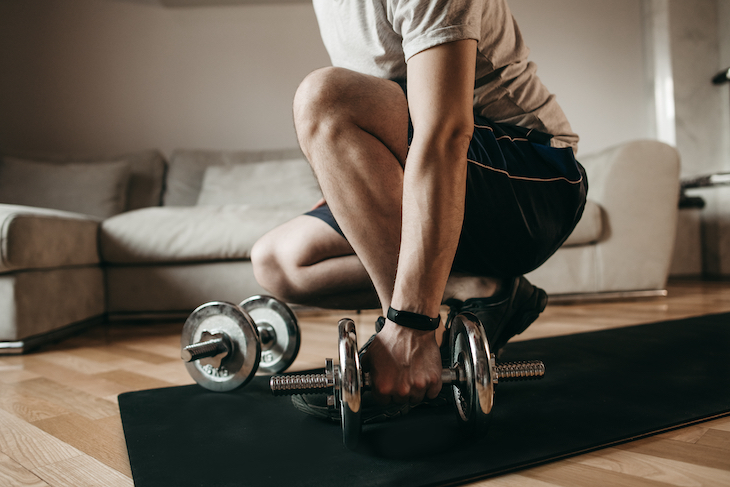 Man engaged with metal dumbbells on black mat at home using wrist smart watch. Sports, technology, health care, body, online training