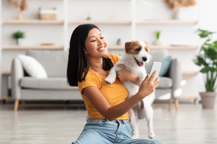 Young woman sitting on floor in living room, taking selfie with cute dog